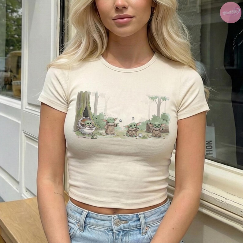 Puede incluir: Camiseta de color crema, de manga corta, con una ilustraci&oacute;n de dibujos animados de Grogu de The Mandalorian. El gr&aacute;fico muestra a Grogu en varias poses en un entorno forestal. La camiseta tiene una textura acanalada.