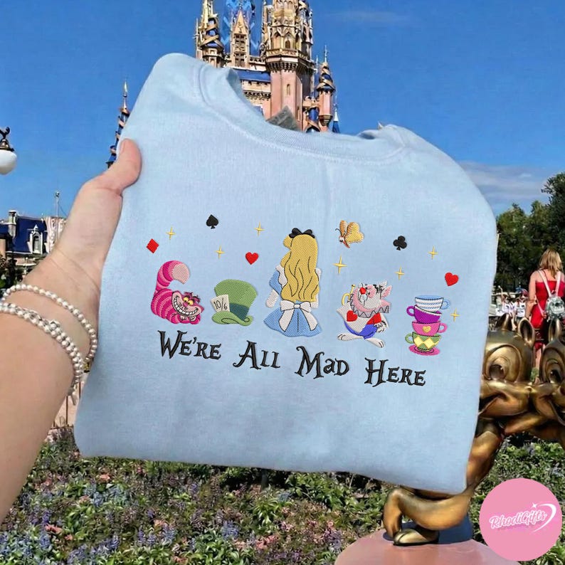 Puede incluir: Sudadera azul claro con un dise&ntilde;o bordado de personajes de Alicia en el pa&iacute;s de las maravillas. El dise&ntilde;o incluye el Gato de Cheshire, el sombrero del Sombrerero Loco, Alicia, el Sombrerero Loco y tazas de t&eacute;. El texto "We're All Mad Here" est&aacute; bordado.