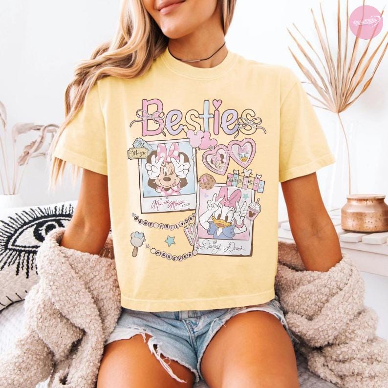 Puede incluir: Camiseta amarilla clara con la palabra "Besties" en una fuente l&uacute;dica. La camiseta presenta ilustraciones de Minnie Mouse y Daisy Duck en marcos estilo polaroid, junto con corazones y elementos decorativos.