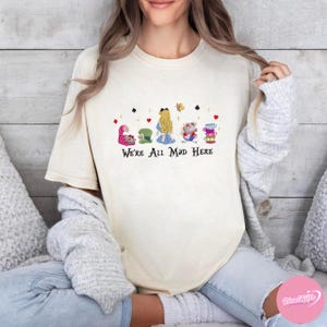 Puede incluir: Camiseta color crema con un gr&aacute;fico colorido de personajes de Alicia en el pa&iacute;s de las maravillas, incluyendo el Gato de Cheshire, el Sombrerero Loco, Alicia y el Conejo Blanco. El texto "We're All Mad Here" est&aacute; impreso debajo.