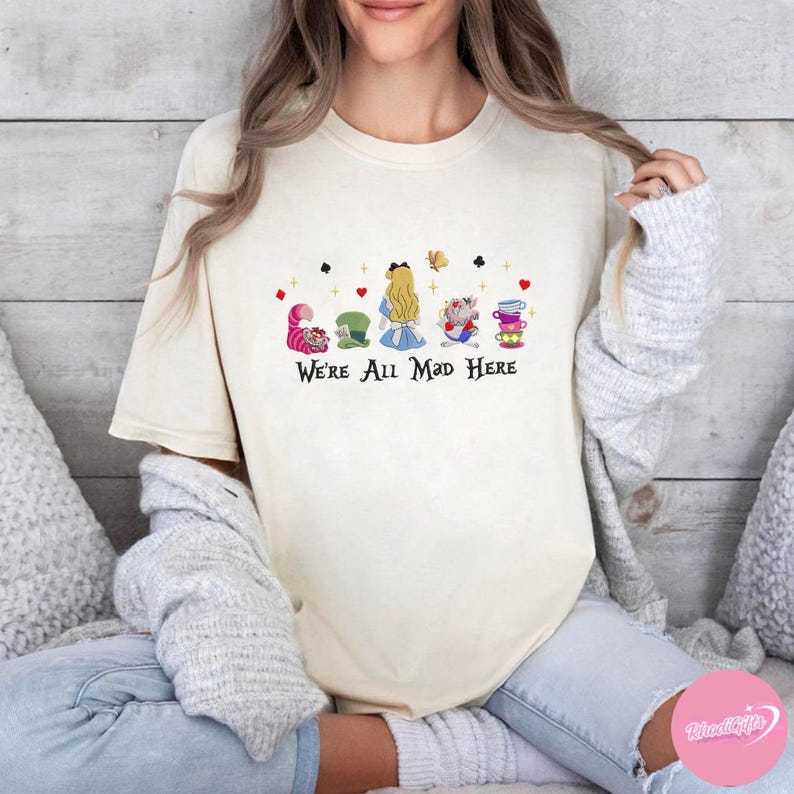 Puede incluir: Camiseta color crema con un gr&aacute;fico colorido de personajes de Alicia en el pa&iacute;s de las maravillas, incluyendo el Gato de Cheshire, el Sombrerero Loco, Alicia y el Conejo Blanco. El texto "We're All Mad Here" est&aacute; impreso debajo.