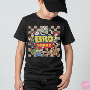May include: Black t-shirt featuring a colorful graphic of characters from the animated movie Toy Story. The design includes the text "BRO STORY" in yellow and blue, with a checkered background. The shirt is worn by a person.