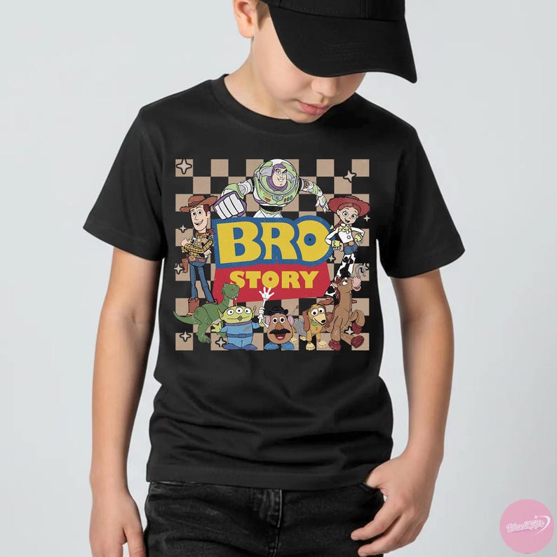 May include: Black t-shirt featuring a colorful graphic of characters from the animated movie Toy Story. The design includes the text "BRO STORY" in yellow and blue, with a checkered background. The shirt is worn by a person.