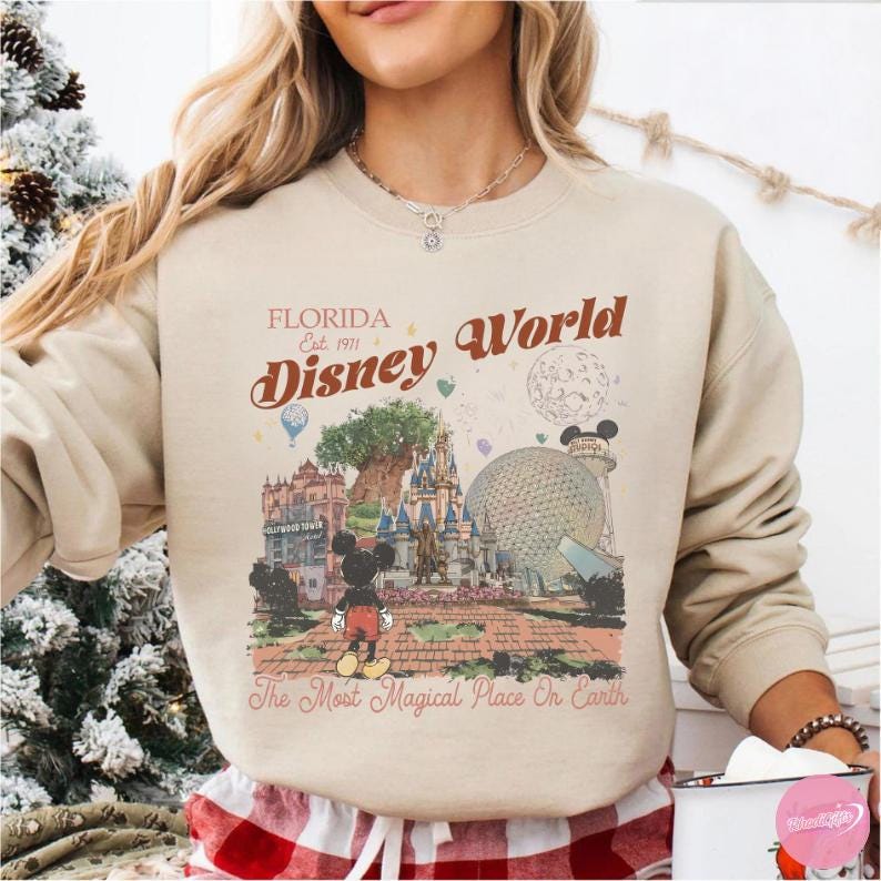 May include: Beige sweatshirt featuring a colorful graphic of Disney World in Florida. The design includes Mickey Mouse, iconic park landmarks, and the text "Florida Disney World Est. 1971" and "The Most Magical Place On Earth."