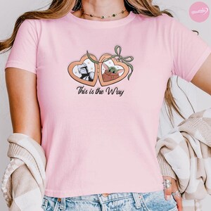 K&ouml;nnte beinhalten: Hellrosa T-Shirt mit einem herzf&ouml;rmigen Design, das eine Figur mit Helm und einen gr&uuml;nen Au&szlig;erirdischen zeigt. Der Text "This is the Way" ist darunter gedruckt. Das T-Shirt hat kurze &Auml;rmel.