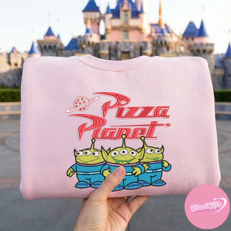 May include: A light pink sweatshirt featuring the Pizza Planet logo and three green alien characters in blue uniforms. The red logo includes a planet with a pizza slice. The sweatshirt is a crew neck style.