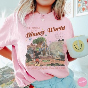 May include: A light pink t-shirt featuring a Disney World graphic with Mickey Mouse, a castle, and the text "Florida Disney World Est. 1971." The shirt also has the phrase "The Most Magical Place On Earth." A white mug with a yellow smiley face is held in the image.