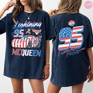May include: Navy blue t-shirt featuring a graphic of Lightning McQueen from the Cars movie. The front design includes the text "Lightning 95 McQueen" with a patriotic color scheme. The back of the shirt displays a large "95" with the text "Lightning McQueen".