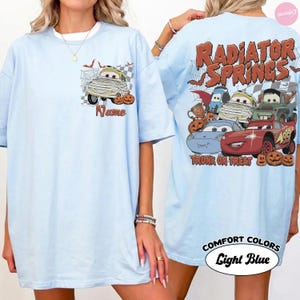 May include: A light blue t-shirt featuring a small graphic on the front chest of a mummy car with the word Name. The back displays a large graphic of multiple cartoon cars with the text Radiator Springs Trunk or Treat and pumpkins.