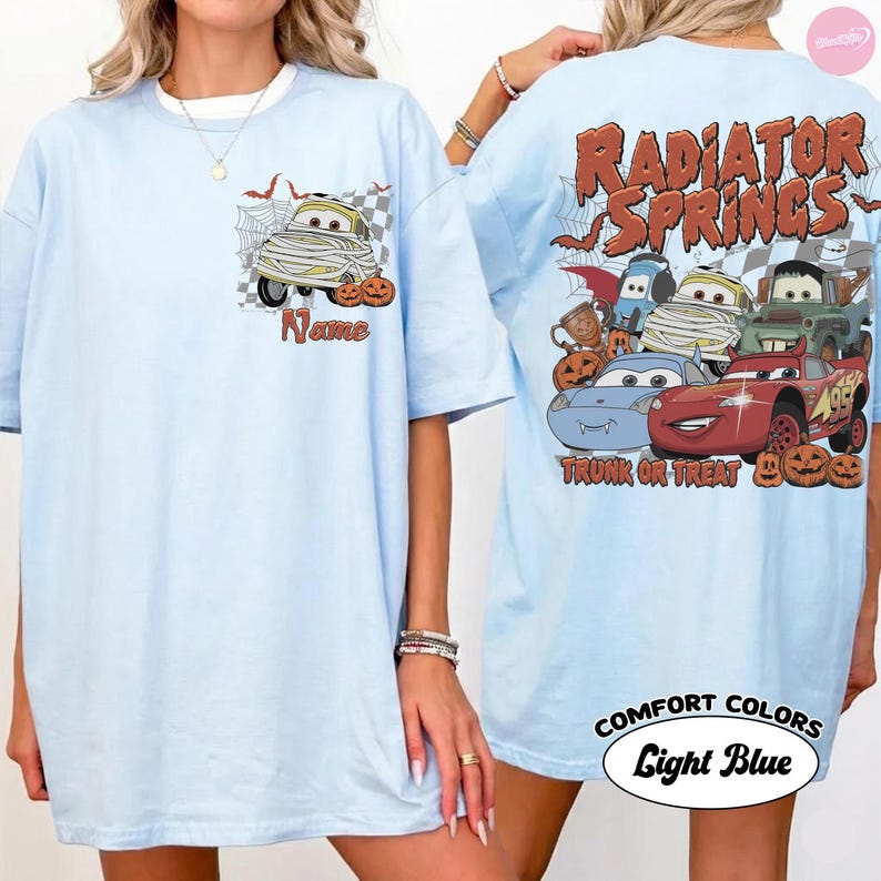 May include: A light blue t-shirt featuring a small graphic on the front chest of a mummy car with the word Name. The back displays a large graphic of multiple cartoon cars with the text Radiator Springs Trunk or Treat and pumpkins.