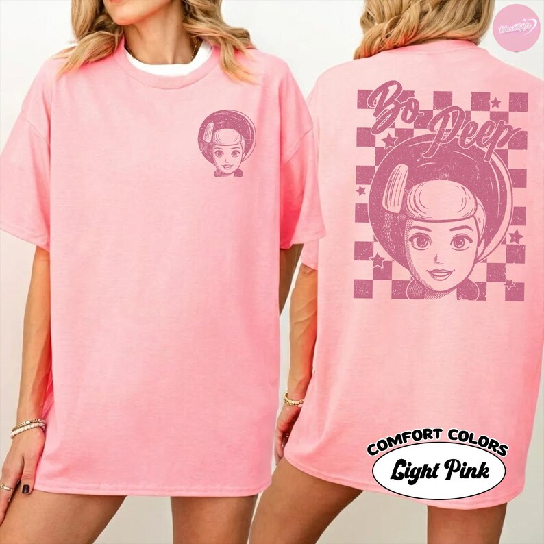 以下が含まれることがあります： トイ・ストーリーのボー・ピープのグラフィックが特徴のライトピンクのTシャツ。前面にはボー・ピープの小さな画像があり、背面には「Bo Peep」の文字とチェック柄の背景が表示されています。シャツには「Comfort Colors Light Pink」と表示されています。