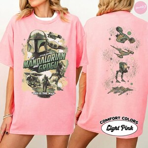 Puede incluir: Camiseta rosa claro con un dise&ntilde;o de Star Wars con The Mandalorian y Grogu. El gr&aacute;fico frontal incluye el casco mandaloriano, Grogu y varios personajes. La parte posterior de la camiseta tiene im&aacute;genes adicionales de Star Wars. El texto "THIS IS THE WAY" tambi&eacute;n es visible.