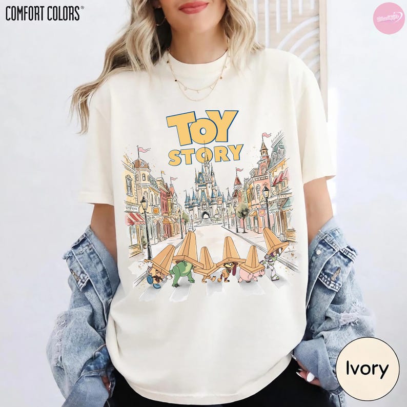 May include: Ivory-colored Comfort Colors t-shirt featuring the words "TOY STORY" in yellow and blue. The graphic depicts a cartoon scene with toy characters and a castle. The shirt is a relaxed fit.