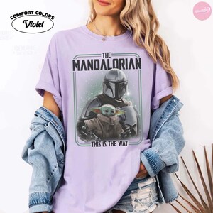 K&ouml;nnte beinhalten: Violettes Comfort Colors T-Shirt mit einem Aufdruck von The Mandalorian und Grogu. Das Design enth&auml;lt den Text "THE MANDALORIAN" und "THIS IS THE WAY". Das Shirt hat eine helle violette Farbe.