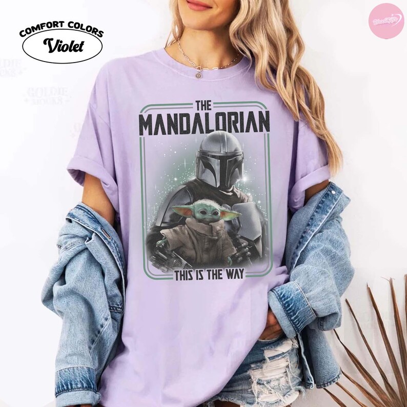 K&ouml;nnte beinhalten: Violettes Comfort Colors T-Shirt mit einem Aufdruck von The Mandalorian und Grogu. Das Design enth&auml;lt den Text "THE MANDALORIAN" und "THIS IS THE WAY". Das Shirt hat eine helle violette Farbe.