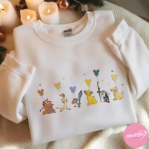 K&ouml;nnte beinhalten: Wei&szlig;es Sweatshirt mit Rundhalsausschnitt und einem farbenfrohen Aufdruck von Figuren aus Der K&ouml;nig der L&ouml;wen, die Mickey Mouse-f&ouml;rmige Ballons halten. Das Design umfasst Simba, Timon, Pumbaa, Zazu und andere Charaktere. Goldene Sterne sind verstreut.