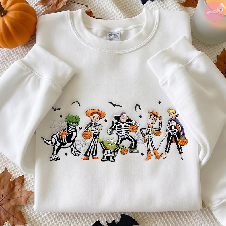 May include: A white crewneck sweatshirt featuring a graphic print of six animated characters dressed in skeleton costumes. The characters are holding orange jack-o'-lanterns, surrounded by small black bats and spider web illustrations.