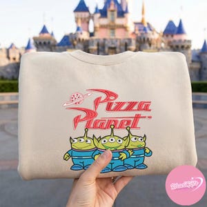 May include: Beige sweatshirt featuring the Pizza Planet logo in red and three green alien characters in blue uniforms. The sweatshirt has a crew neck and long sleeves. The background shows a castle.