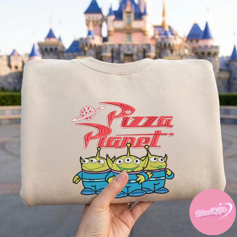 May include: Beige sweatshirt featuring the Pizza Planet logo in red and three green alien characters in blue uniforms. The sweatshirt has a crew neck and long sleeves. The background shows a castle.
