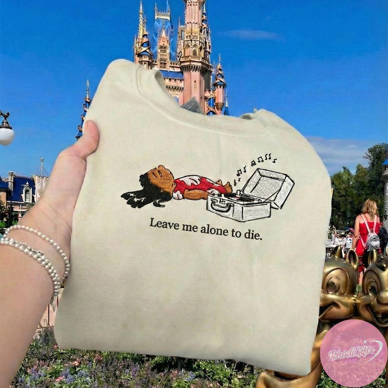 May include: Cream-colored sweatshirt featuring a cartoon character lying next to a record player. The character is wearing a red and white outfit. The text "Leave me alone to die" is printed below the graphic.