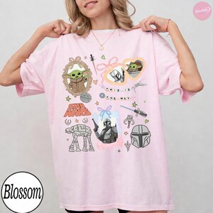 May include: Light pink t-shirt featuring Star Wars graphics. The design includes images of Grogu, the Mandalorian, and the text "This is the Way." Other graphics include an AT-AT walker, a helmet, and various Star Wars-themed elements.
