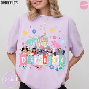 Pu&ograve; includere: T-shirt Orchid Comfort Colors con grafica Disneyland. Il design include personaggi dei cartoni animati, un castello e la scritta "Disneyland" in lettere colorate. Elementi aggiuntivi includono palme, fiori e una tavola da surf.