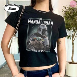 K&ouml;nnte beinhalten: Schwarzes T-Shirt mit einer Grafik von The Mandalorian und Grogu, mit dem Text "THE MANDALORIAN" und "THIS IS THE WAY." Das Shirt besteht aus Comfort Colors-Stoff.