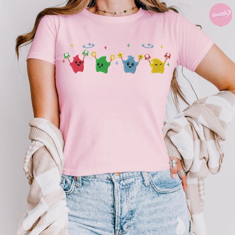 Puede incluir: Camiseta rosa claro con un dise&ntilde;o de dibujos animados. El dise&ntilde;o incluye cuatro personajes en forma de estrella en rojo, verde, azul y amarillo, cada uno sosteniendo una estrella y una seta. La camiseta es de material suave.