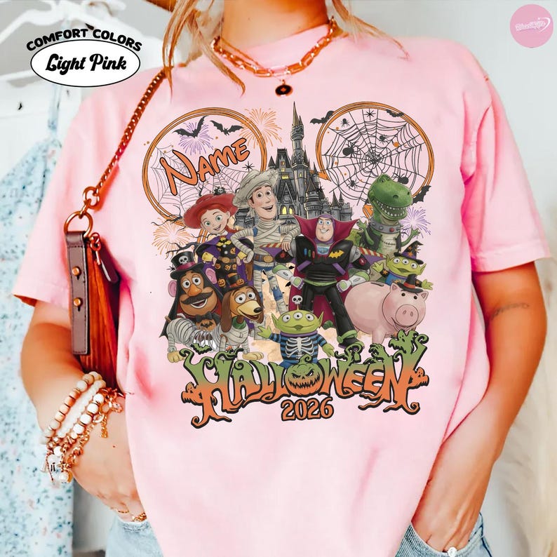 May include: A light pink short-sleeve t-shirt featuring a graphic of various animated characters in Halloween costumes, including a cowboy, space ranger, and dinosaur. The design includes a castle, spiderwebs, and the text Name, Halloween, and 2026.