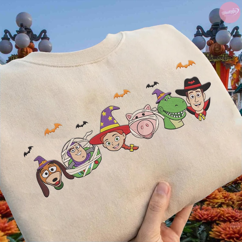 May include: Cream-colored sweatshirt featuring embroidered Halloween-themed Toy Story characters. The design includes Slinky Dog, Buzz Lightyear, Jessie, a pig, Rex, and Woody, each in a Halloween costume, with orange bat accents.