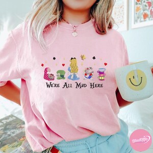 Puede incluir: Camiseta rosa claro con un dise&ntilde;o de Alicia en el pa&iacute;s de las maravillas. El gr&aacute;fico incluye personajes como el Gato de Cheshire, el Sombrerero Loco, Alicia y las tazas de t&eacute;, con el texto "We're All Mad Here". Tambi&eacute;n se ve una taza blanca con una carita sonriente amarilla.