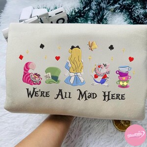 Puede incluir: Coj&iacute;n bordado de color crema con personajes de Alicia en el pa&iacute;s de las maravillas. El dise&ntilde;o incluye a Alicia, el Gato de Cheshire, el sombrero del Sombrerero Loco y tazas apiladas, con el texto "We're All Mad Here".