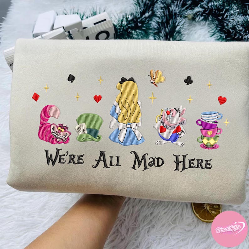 Puede incluir: Coj&iacute;n bordado de color crema con personajes de Alicia en el pa&iacute;s de las maravillas. El dise&ntilde;o incluye a Alicia, el Gato de Cheshire, el sombrero del Sombrerero Loco y tazas apiladas, con el texto "We're All Mad Here".