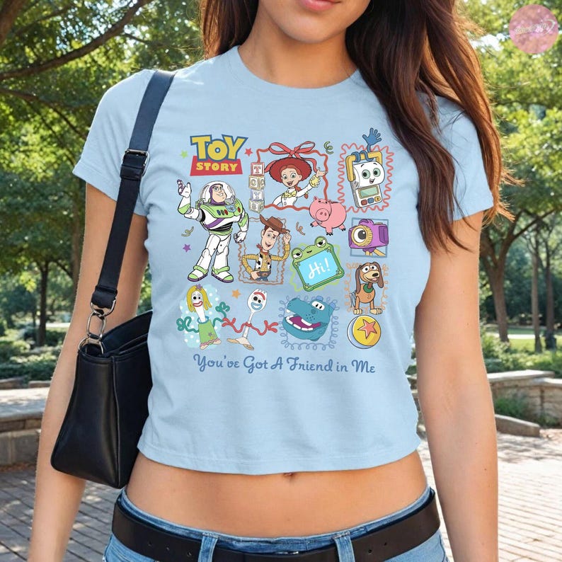 May include: A light blue short-sleeved t-shirt featuring a colorful Toy Story graphic print with characters including Buzz Lightyear, Woody, Jessie, and Forky, along with the text "Toy Story" and "You've Got A Friend in Me" in a playful font.