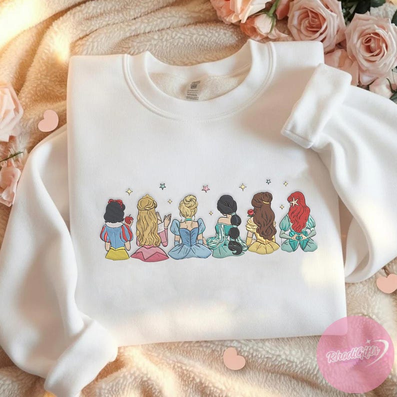 May include: White crewneck sweatshirt featuring an embroidered design of seven cartoon princesses in various dresses and hairstyles. The design includes Snow White, Cinderella, and Ariel, among others, with star accents above them. The sweatshirt is a casual, comfortable style.