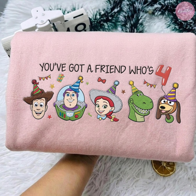 K&ouml;nnte beinhalten: Ein hellrosa Sweatshirt mit dem Text "YOU'VE GOT A FRIEND WHO'S 4" und Cartoon-Charakter-Illustrationen. Das Design zeigt Woody, Buzz Lightyear, Jessie, Rex und Slinky Dog, die alle eine Partyh&uuml;te tragen. Ein roter Ballon mit der Zahl 4 ist ebenfalls enthalten.