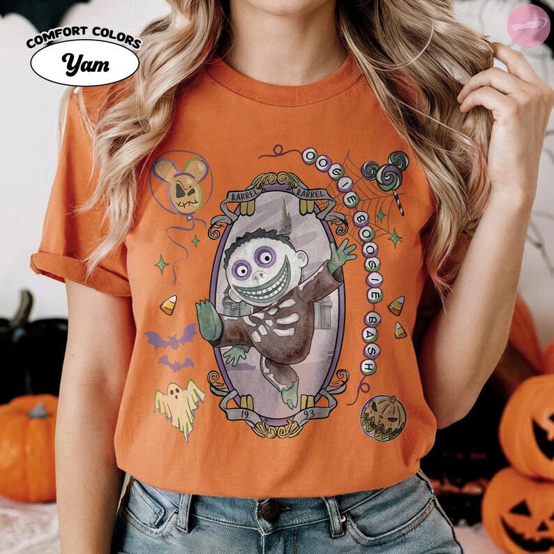 May include: An orange short-sleeve t-shirt featuring a graphic of the character Barrel from The Nightmare Before Christmas inside an ornate oval frame. The design includes the text Oogie Boogie Bash, a ghost, bats, candy corn, and a balloon.