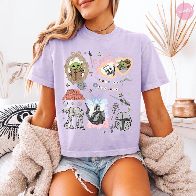 May include: A light purple t-shirt featuring various Star Wars-themed illustrations. The design includes images of Grogu, the Mandalorian, and other iconic elements. The phrase "THIS IS THE WAY" is also printed on the shirt.