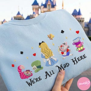 Puede incluir: Sudadera azul claro con bordados de personajes de Alicia en el Pa&iacute;s de las Maravillas, incluyendo a Alicia, el Gato de Cheshire y el Sombrerero Loco. La frase "We're All Mad Here" est&aacute; bordada en negro. El dise&ntilde;o incluye corazones, estrellas y s&iacute;mbolos de naipes.
