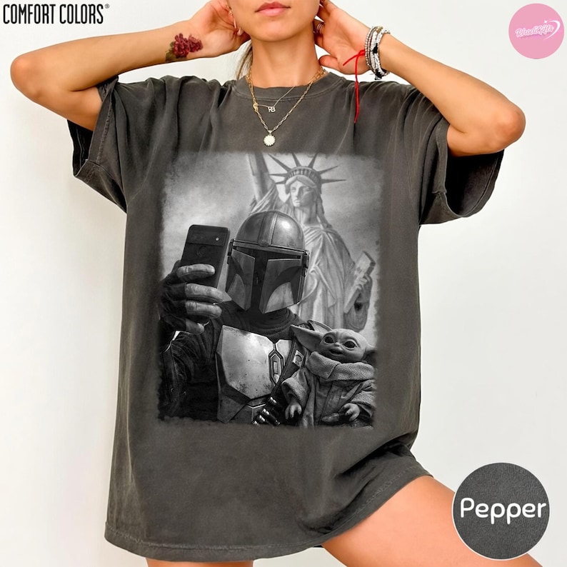 May include: Dark grey Comfort Colors t-shirt featuring a black and white graphic of The Mandalorian taking a selfie with Grogu and the Statue of Liberty in the background. The shirt is labeled "Pepper".