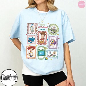 May include: A light blue t-shirt featuring a graphic print of various Toy Story characters arranged in colorful postage stamp-style frames. The text "You've Got A Friend In Me" is printed in the center, surrounded by illustrations of Woody, Buzz Lightyear, Jessie, and other characters.