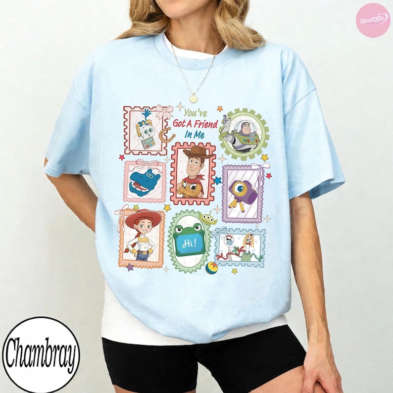 May include: A light blue t-shirt featuring a graphic print of various Toy Story characters arranged in colorful postage stamp-style frames. The text "You've Got A Friend In Me" is printed in the center, surrounded by illustrations of Woody, Buzz Lightyear, Jessie, and other characters.