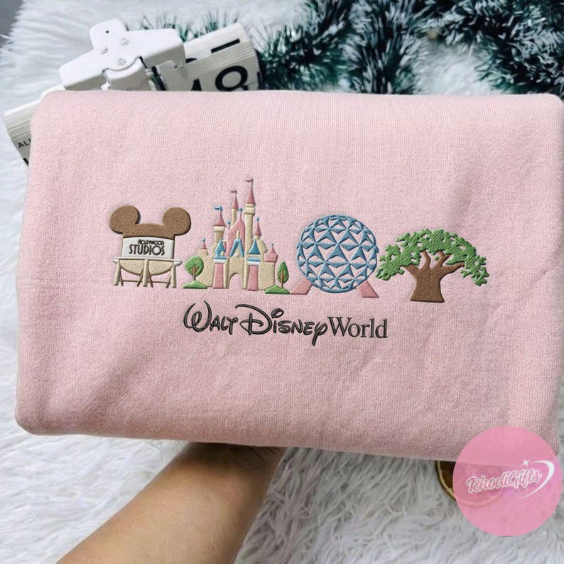 Peut inclure: Un sweat-shirt rose clair avec un motif brod&eacute; repr&eacute;sentant le logo Walt Disney World et des monuments embl&eacute;matiques. Le motif comprend le logo Hollywood Studios, un ch&acirc;teau, une sph&egrave;re et un arbre. Le texte "Walt Disney World" est brod&eacute; en dessous.