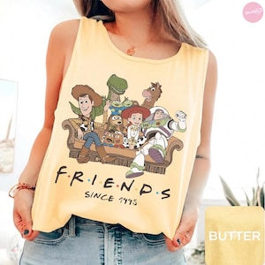 May include: A pale yellow sleeveless tank top featuring a graphic print of Toy Story characters sitting on a brown sofa. Below the characters, the text reads F R I E N D S SINCE 1995 in black lettering with colorful dots between the letters.