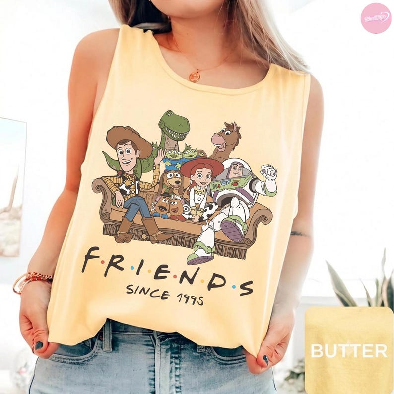 May include: A pale yellow sleeveless tank top featuring a graphic print of Toy Story characters sitting on a brown sofa. Below the characters, the text reads F R I E N D S SINCE 1995 in black lettering with colorful dots between the letters.