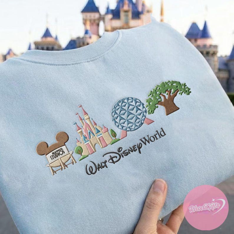 Peut inclure: Sweat-shirt bleu clair avec broderie d'ic&ocirc;nes de Disney World : ch&acirc;teau, Spaceship Earth et arbre. Le texte "Walt Disney World" est brod&eacute; en dessous. Comprend &eacute;galement une t&ecirc;te de Mickey Mouse avec le texte "Hollywood Studios".