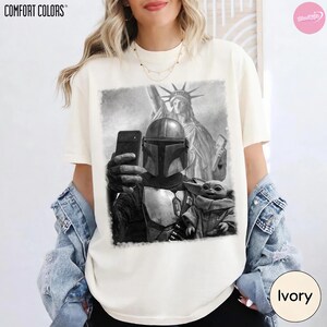 May include: Ivory-colored t-shirt featuring a black and white graphic of a Mandalorian character taking a selfie with Grogu in front of the Statue of Liberty. The shirt has the Comfort Colors logo at the top left.