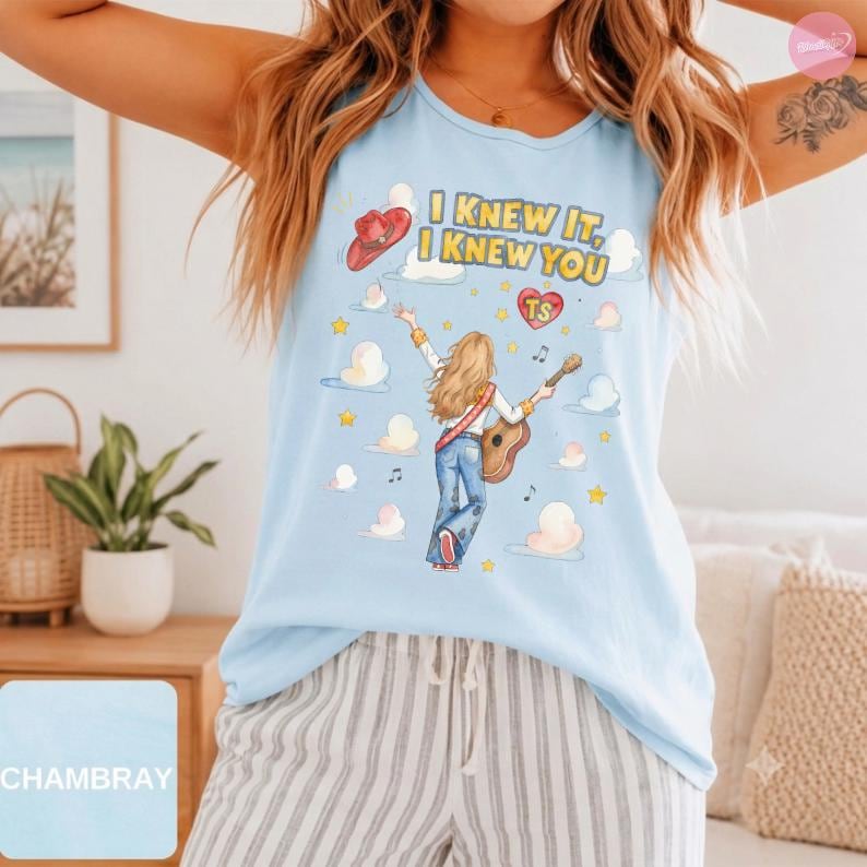 May include: A light blue sleeveless tank top featuring a graphic illustration of a person with long hair playing an acoustic guitar. The text reads I KNEW IT, I KNEW YOU with a red cowboy hat, a heart with TS, musical notes, and yellow stars.
