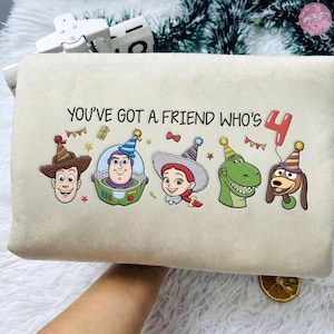 K&ouml;nnte beinhalten: Ein beiges rechteckiges Kissen mit dem Text "YOU'VE GOT A FRIEND WHO'S 4" und Cartoonfiguren aus Toy Story. Die Figuren tragen Partyh&uuml;te und sind von festlichen Dekorationen umgeben.