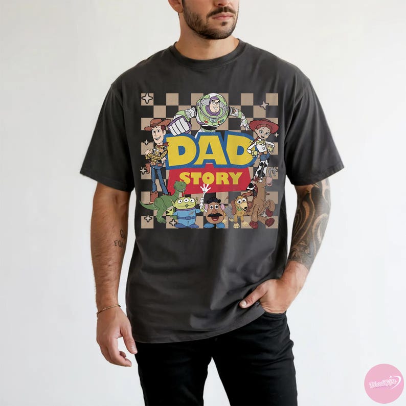 May include: Dark gray t-shirt featuring a colorful graphic of characters from the Toy Story movie, with the text "DAD STORY" in yellow and red. The shirt has a crew neck and short sleeves.
